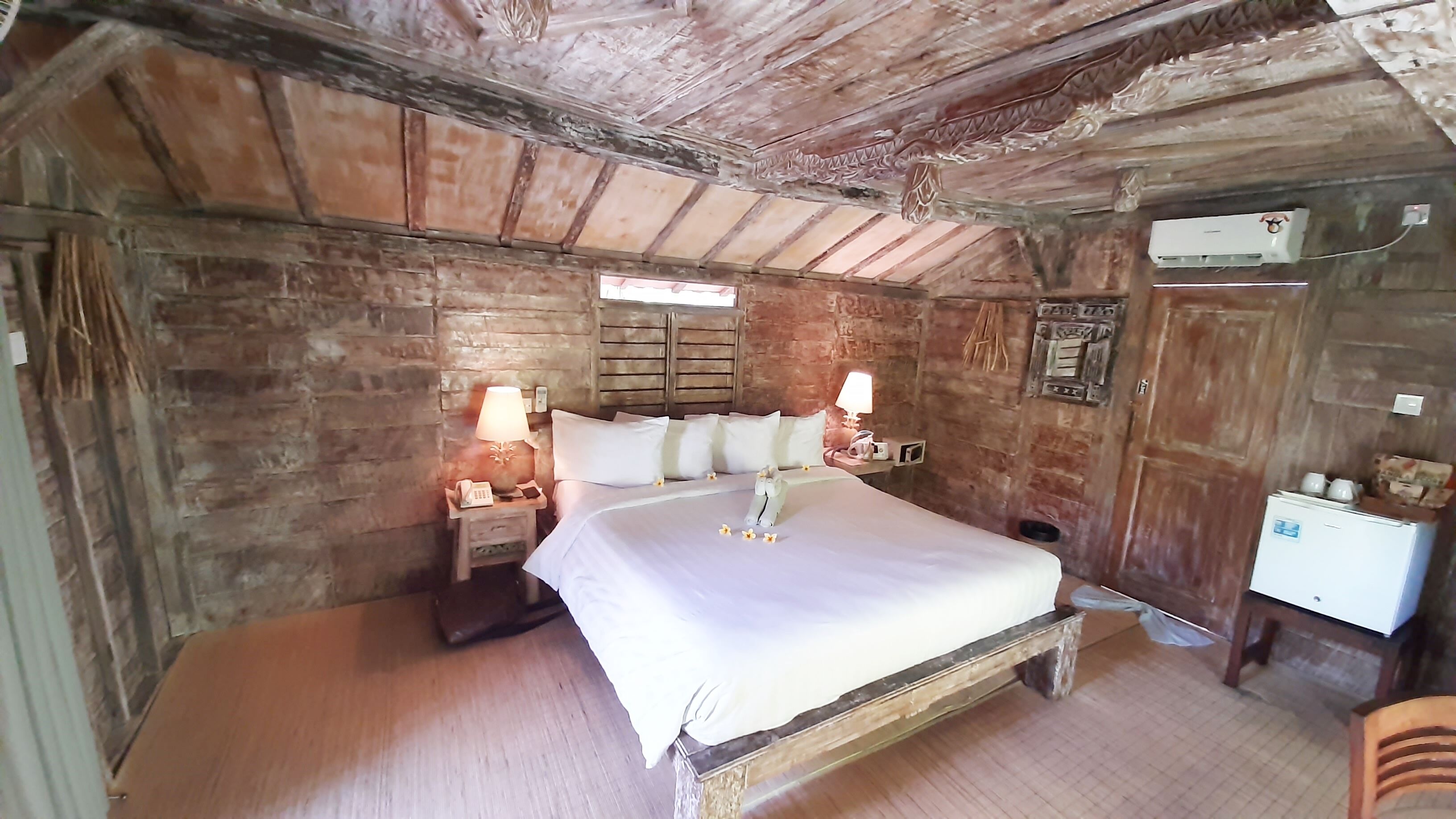 A rustic bedroom with a wooden bed, two bedside tables with lamps, a brick wall, and a small white dresser.
