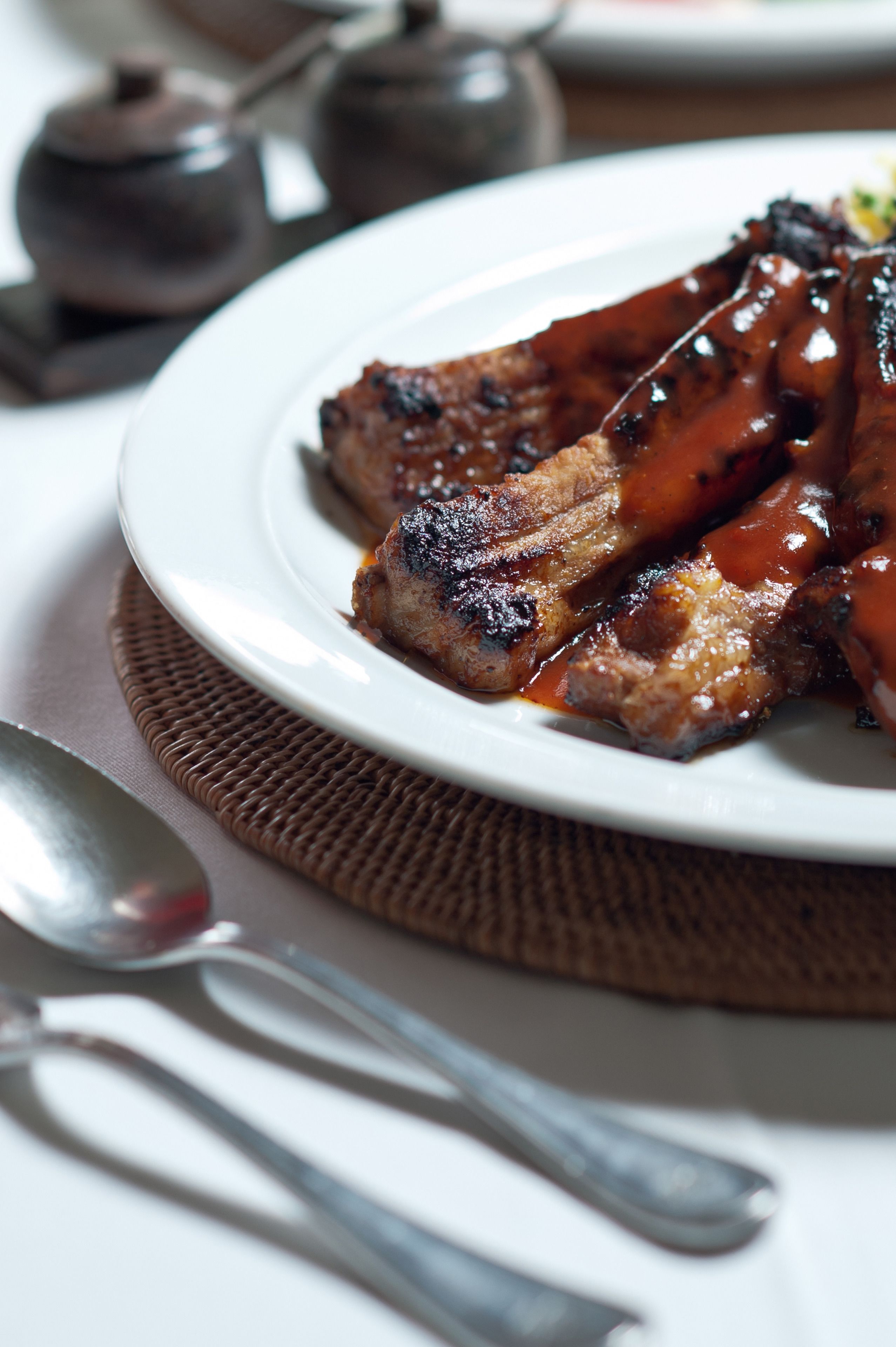 A plate of grilled meat with a glossy sauce, accompanied by a spoon and a teapot on a table.