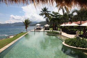 Piscina con vistas al mar y montañas, rodeada de palmeras y sillas de descanso.