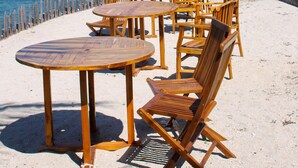 A beachside restaurant with wooden tables and chairs, a view of the ocean, and palm trees. - Sea Breeze Candidasa (Manggis)