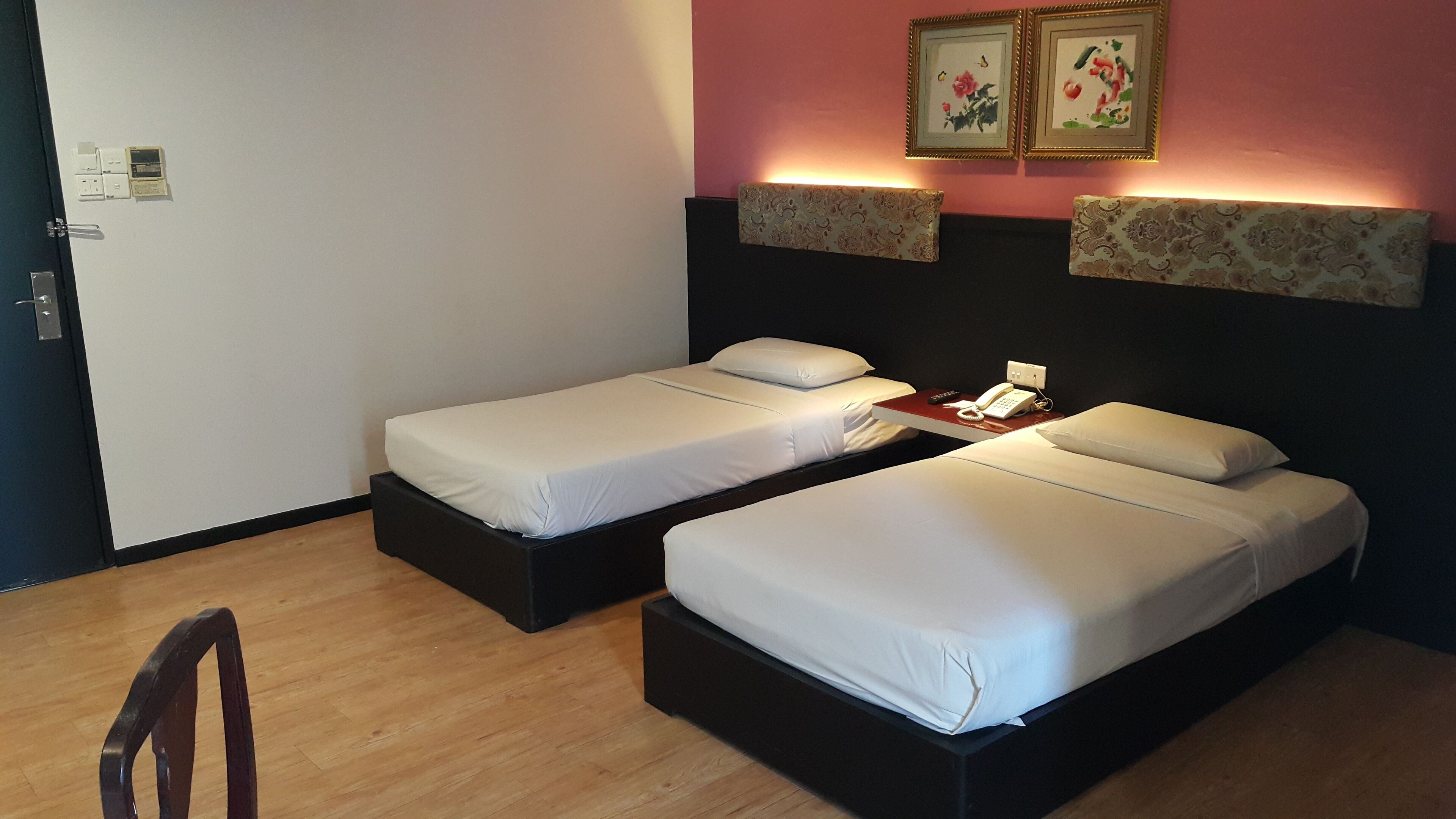 A hotel room with two single beds, a bedside table with a phone and a red object, and a wall-mounted light fixture.