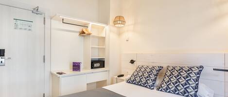 Habitación de hotel con cama, mesita de noche y escritorio con silla. Se observa una estantería en la pared con un abrigo y un objeto morado.