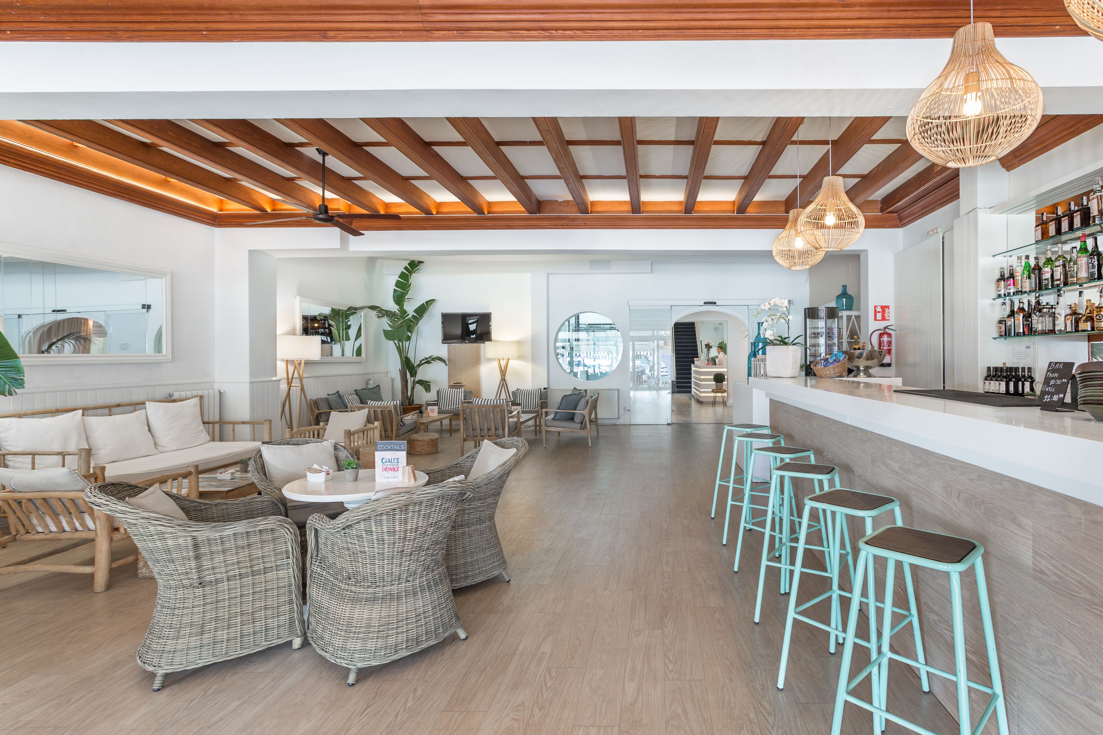 A spacious bar area with wicker furniture and stools, a wooden ceiling, and a bar stocked with bottles.