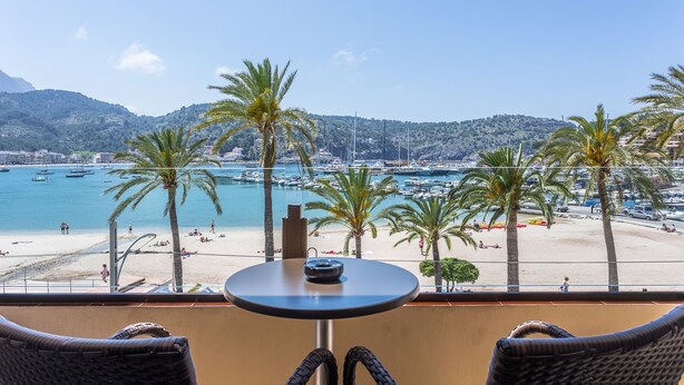Un balcó amb cadires de vimet, una taula petita rodona i vista a una platja amb palmeres i vaixells.