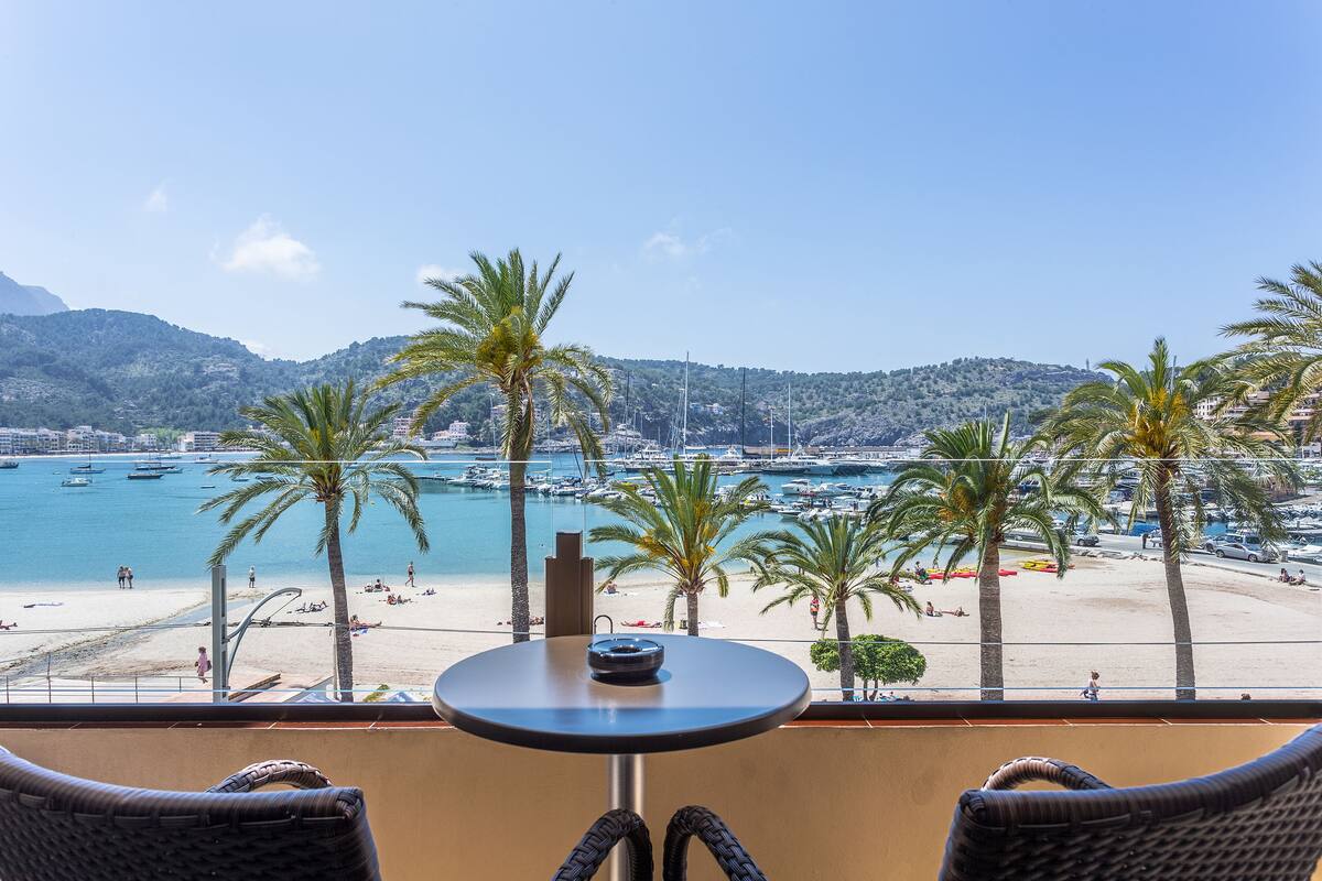 Ein Balkon mit Korbstühlen, einem kleinen runden Tisch und Blick auf einen Strand mit Palmen und Booten.