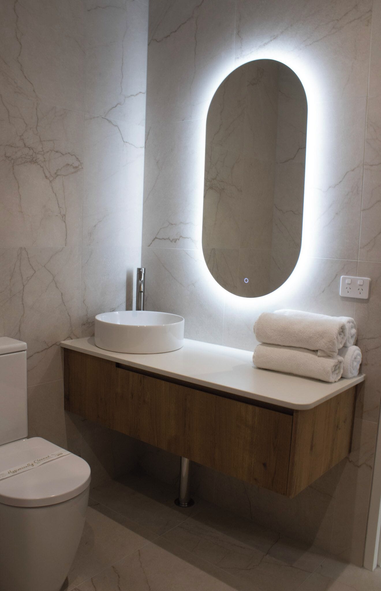 Un baño moderno con tocador de mármol, un espejo redondo con iluminación integrada, inodoro y toallas dobladas con cuidado.