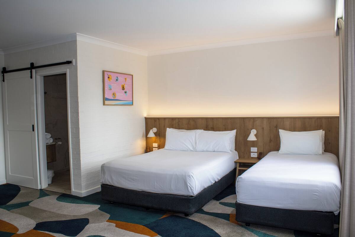 A hotel room with two beds, a wooden headboard, a colorful abstract rug, and a door leading to a bathroom.