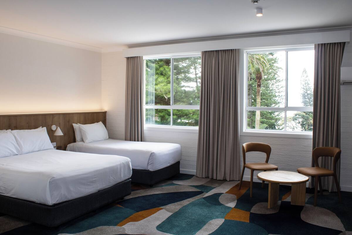 A hotel room with two beds, a small table, two chairs, a window with curtains, and a colorful carpet.