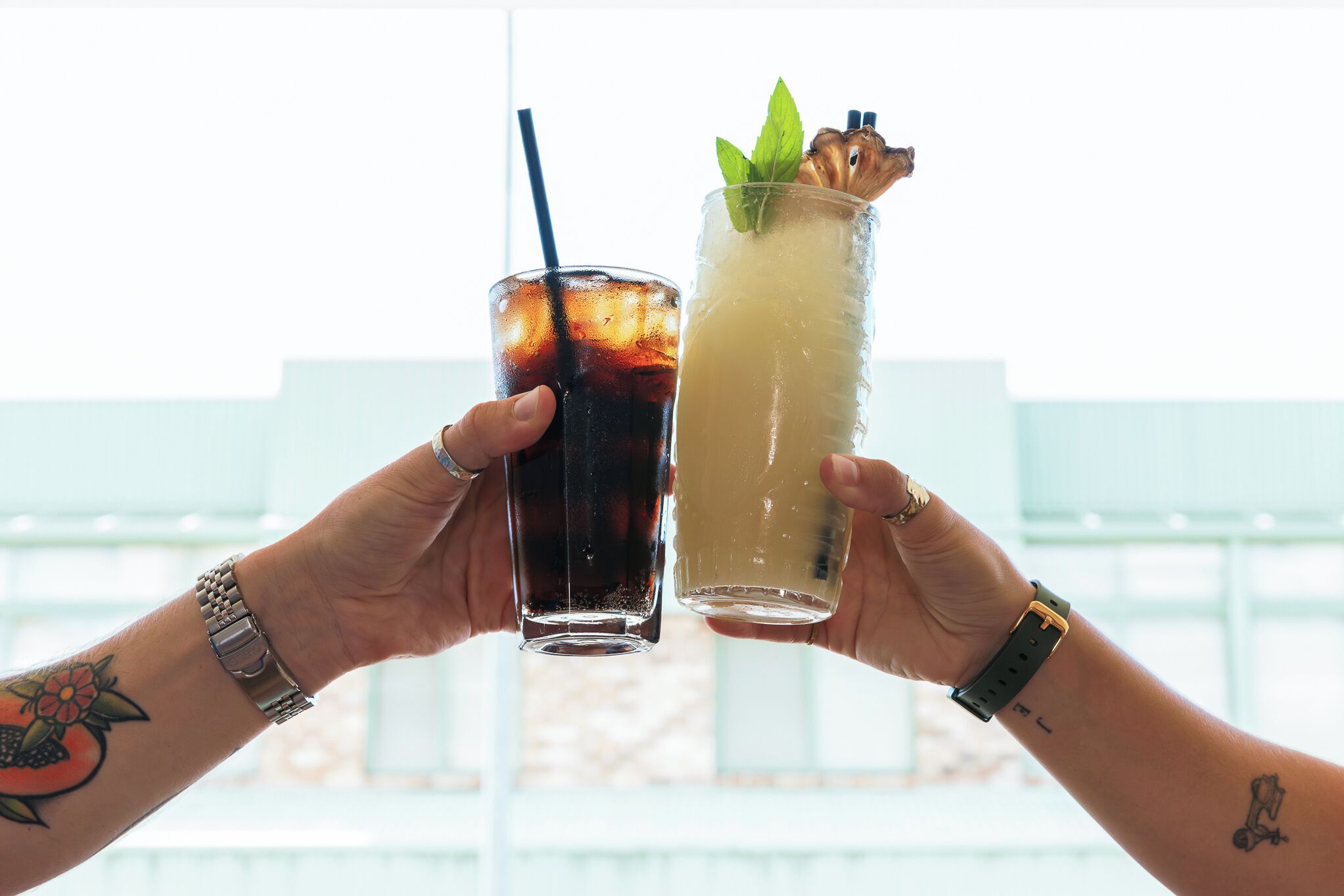 Duas mãos segurando copos com bebidas, um com canudo e enfeite de hortelã, e outro com canudo e gelo.