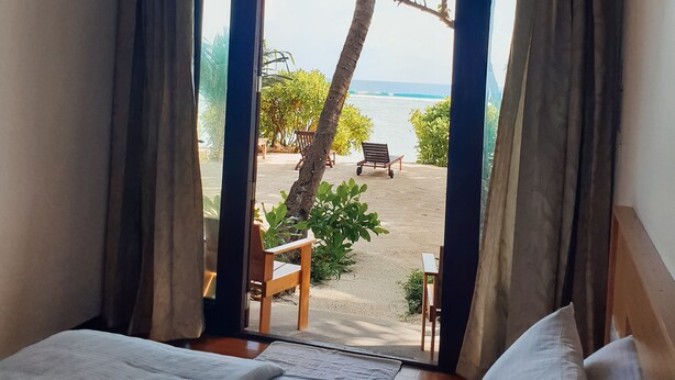 Una habitación con cama, ventana con cortinas y vista a una playa con sillas y una palmera.