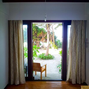A room with a wooden bench, a view of a garden with palm trees and plants, and a door with curtains. - Thundi Sea View  (Fulidhoo)