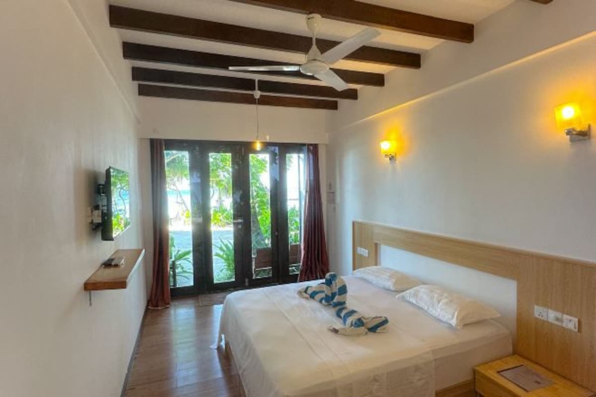 Habitación de hotel con cama, ventilador de techo, vigas de madera, televisión y vistas a la vegetación por la ventana.