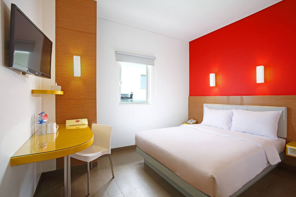 A hotel room with a bed, a desk with a chair, a TV, and a window with blinds.