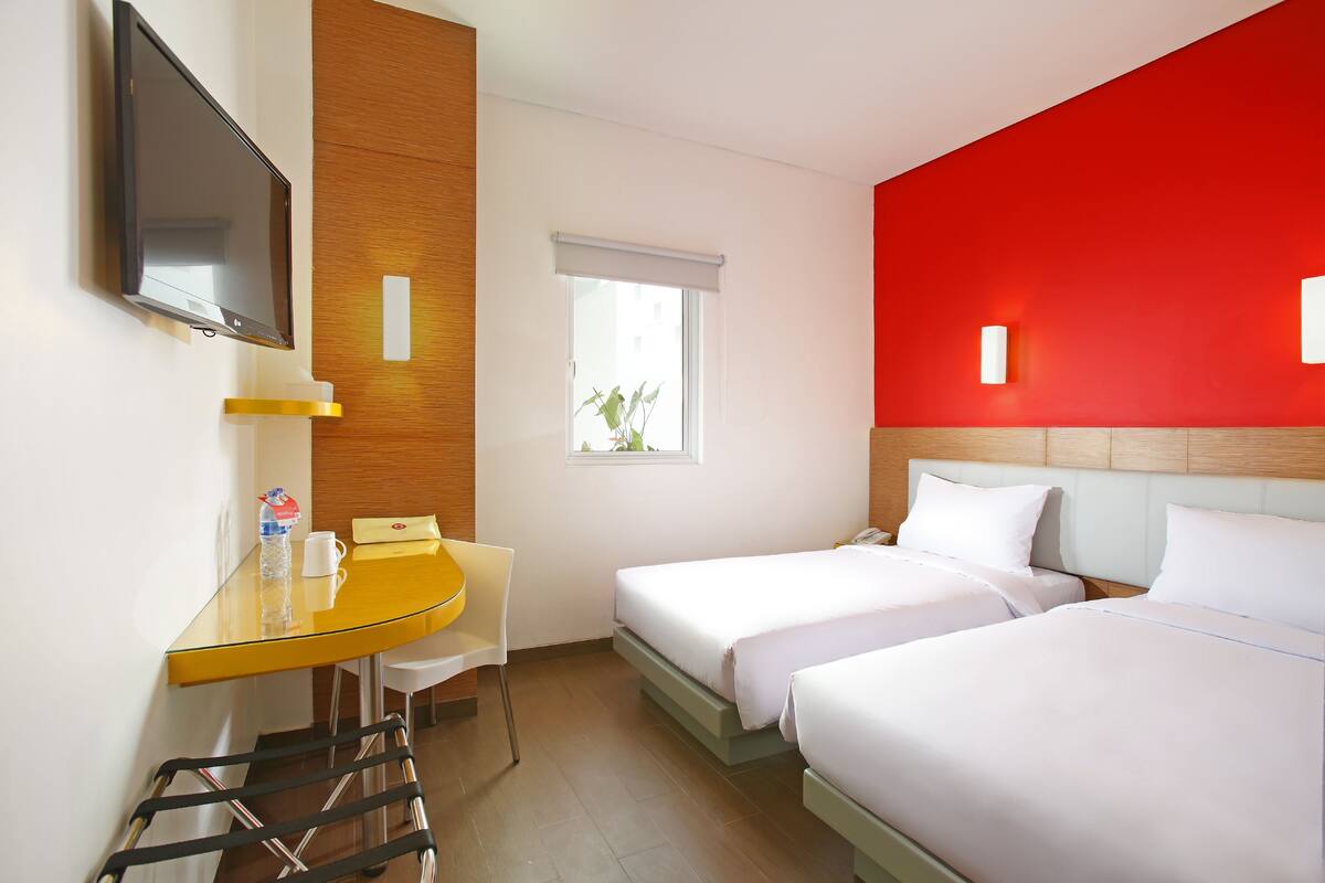 A hotel room with two beds, a desk with a chair, a TV, and a window with a view of greenery.