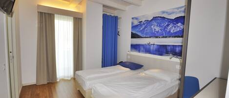 Habitación de hotel con dos camas individuales, un escritorio, una silla, una ventana con cortinas y un televisor montado en la pared.