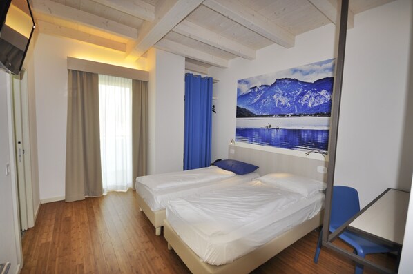 A hotel room with two single beds, a desk, a chair, a window with curtains, and a wall-mounted TV.