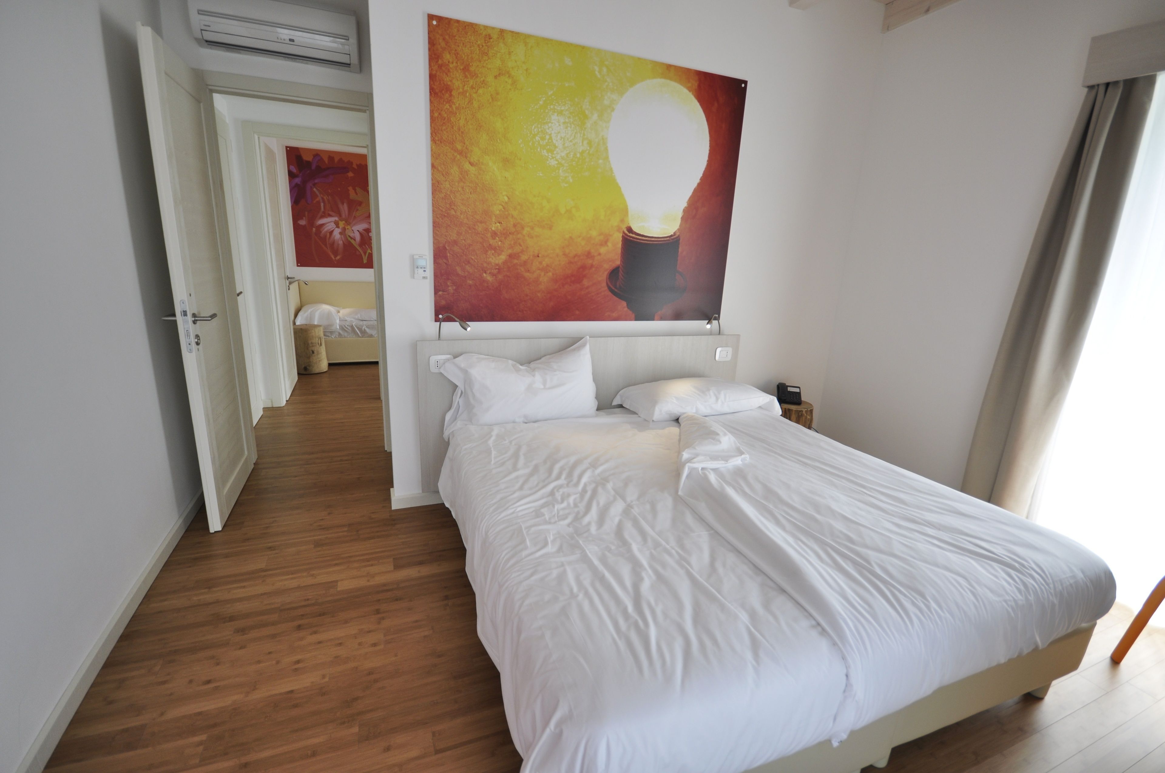 Hotellihuone, jossa on suuri sänky, seinälle kiinnitetty taideteos, jossa on kuva hehkulampusta, puulattia ja ovi, joka johtaa toiseen huoneeseen.