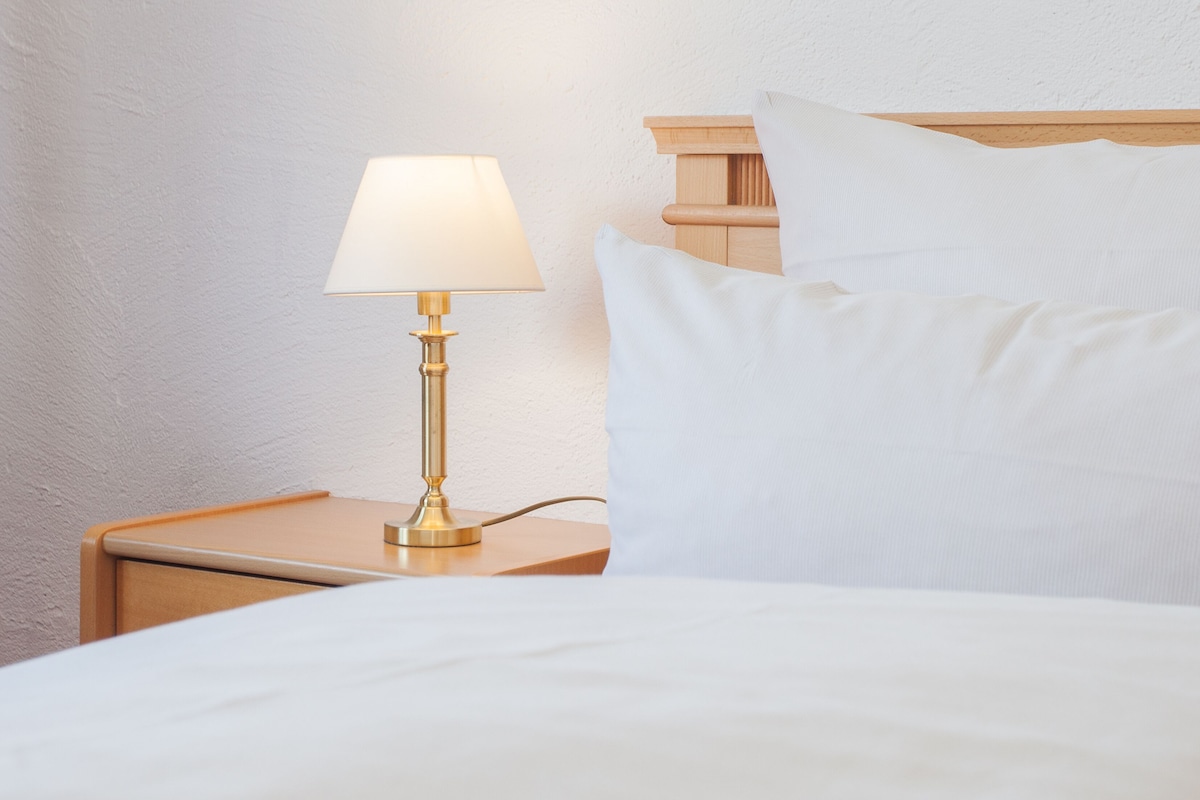 A neatly made bed with white linens, a bedside lamp, and a wooden headboard.