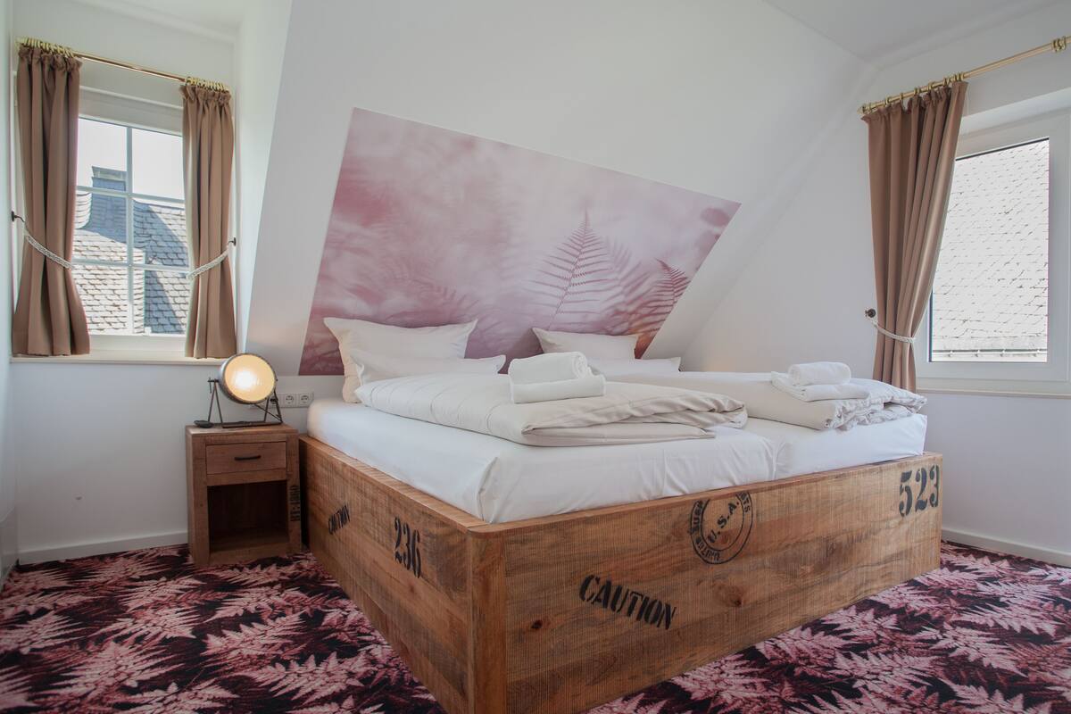 A bedroom with a large bed, a bedside table with a lamp, and a window with curtains.