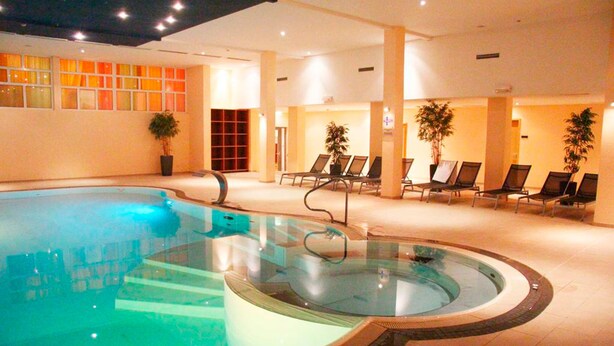 Une grande piscine intérieure dotée d’un mur en verre, entourée de chaises longues et de plantes en pot.
