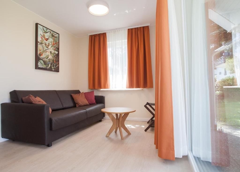 Ein modernes Wohnzimmer mit einem dunklen Sofa, einem runden Holztisch und einem großen Fenster mit orangefarbenen Vorhängen.