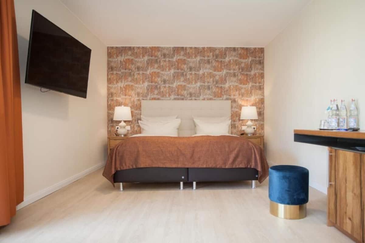 A bedroom with a brick-patterned headboard, a bed with two pillows, bedside lamps, a small round table, a blue ottoman, and a wooden cabinet with bottles on top.
