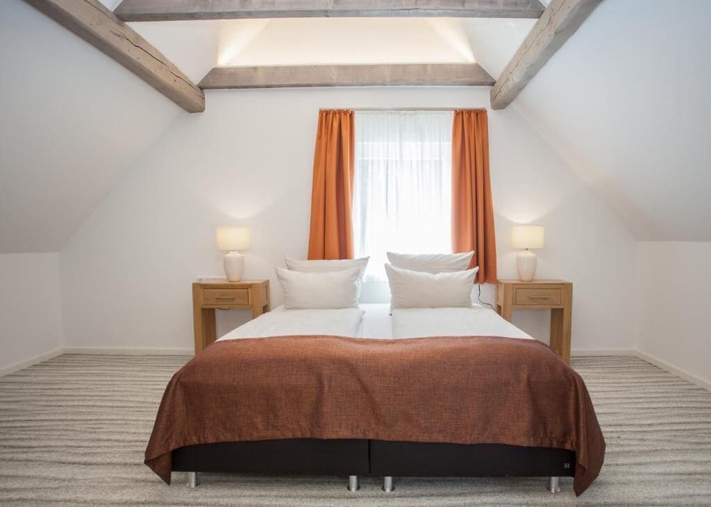 Une chambre avec un grand lit, deux tables de chevet avec des lampes, une fenêtre avec des rideaux orange et un plafond en pente.