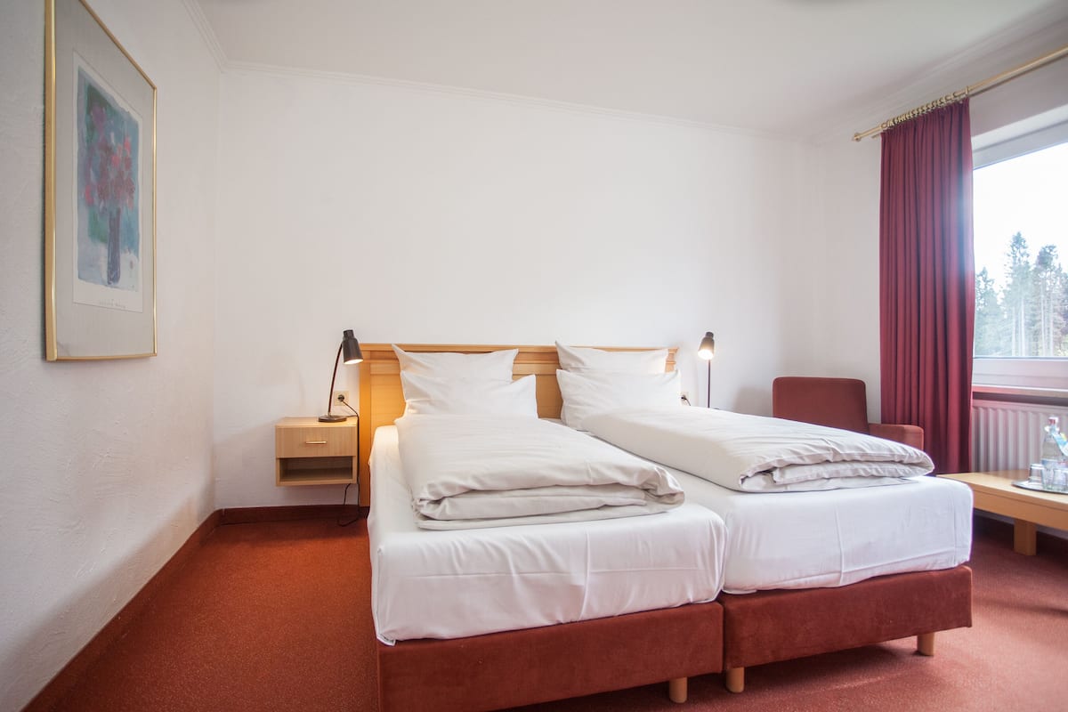 A hotel room with a double bed, a bedside table with a lamp, a chair, a window with curtains, and a painting on the wall.