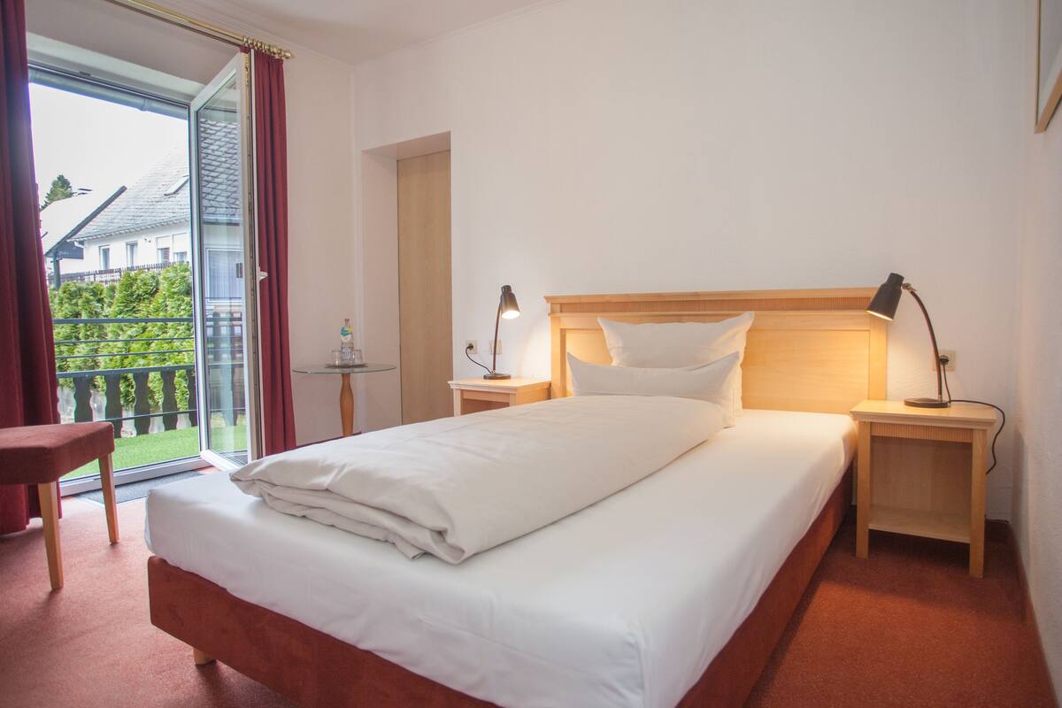 A hotel room with a bed, two bedside tables, a chair, a glass table, and a view of greenery outside.