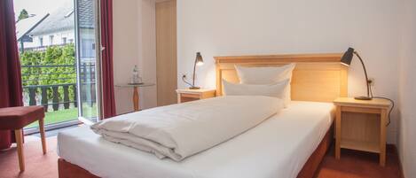 Habitación de hotel con cama, dos mesitas de noche, una silla, una mesita de vidrio y vistas al exterior con vegetación.