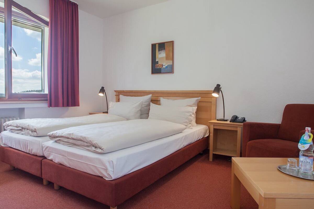 A hotel room with a double bed, a single bed, a red armchair, a wooden nightstand with a lamp, a table with a bottle of water, and a painting on the wall.