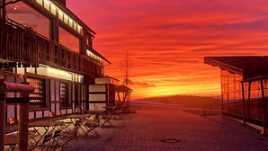 Un bâtiment moderne doté d’une façade en verre, d’un espace extérieur aménagé pour s’asseoir et offrant une vue sur un coucher de soleil se déclinant sur un paysage lointain.
