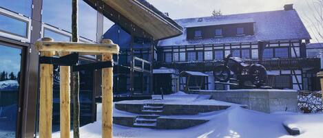 Un espacio exterior cubierto de nieve con una estructura de madera, un edificio con grandes ventanales y un techo nevado.
