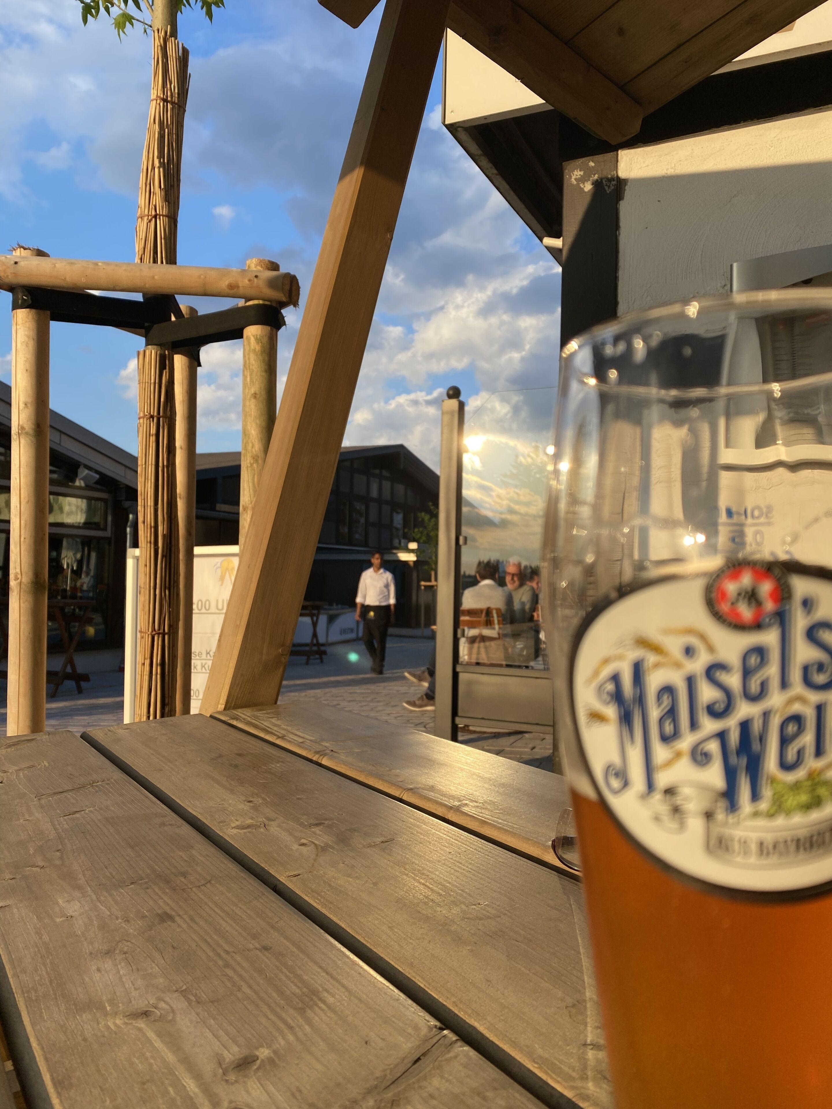 Ein Bierglas mit Etikett, ein Holztisch und im Hintergrund ein Gebäude.