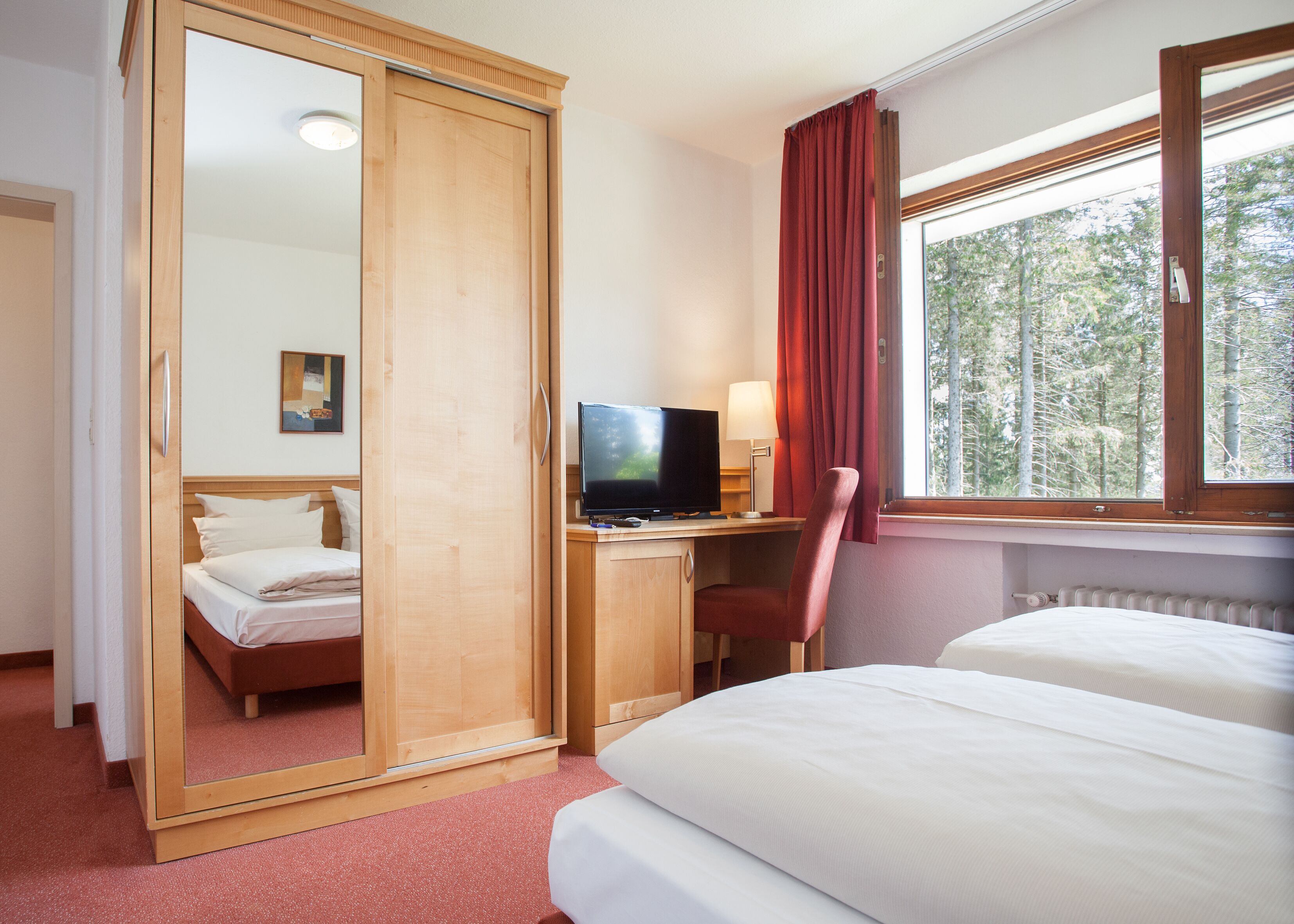 Ein Hotelzimmer mit Bett, Schreibtisch mit Fernseher, Sessel, Fenster mit Vorhängen und Spiegel.