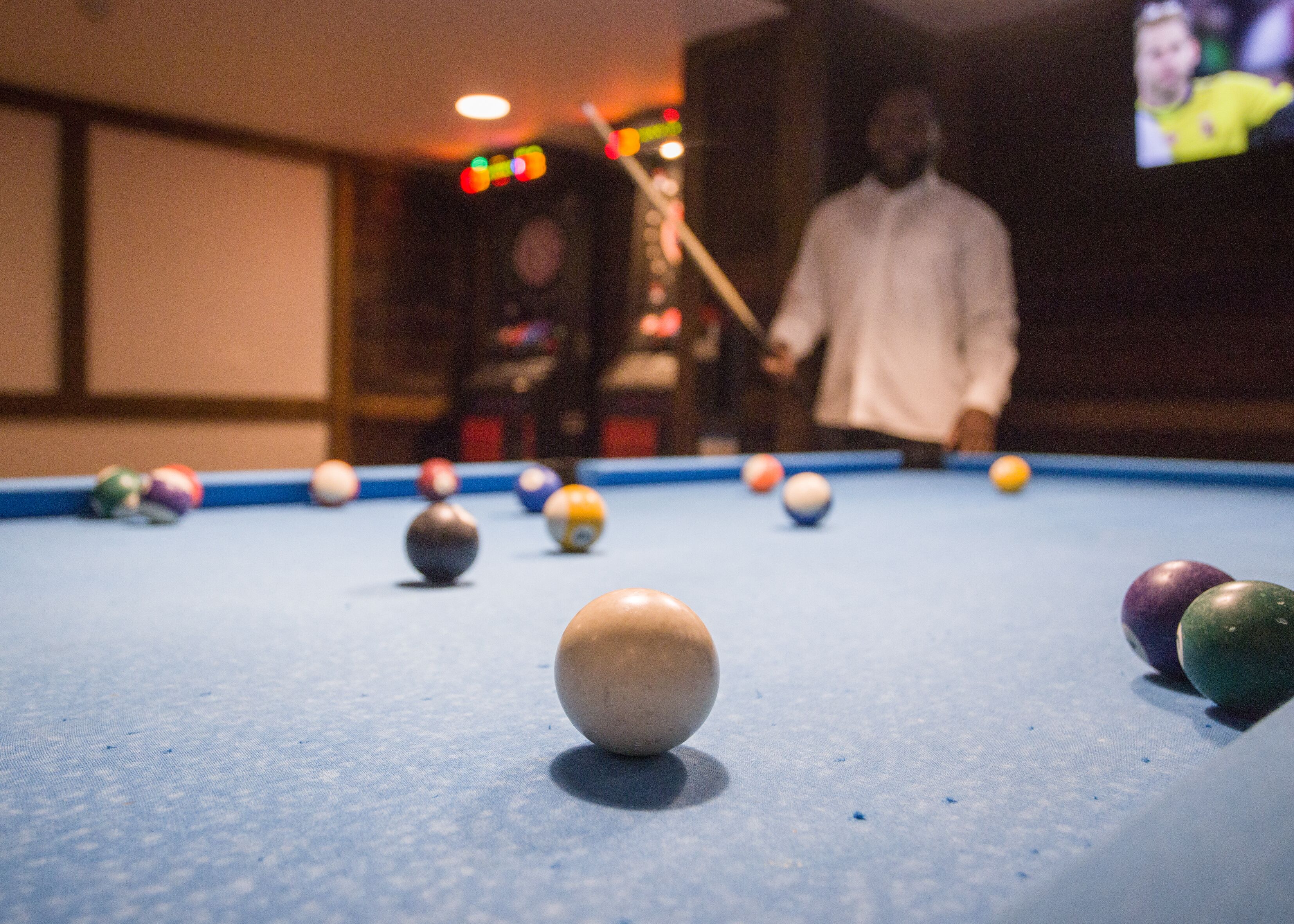 A pool table with balls and a cue stick, a television screen in the background, and a person in the distance.