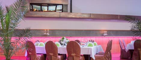 Restaurante com uma mesa comprida posta para refeição, cadeiras com capas estampadas e uma planta decorativa em primeiro plano.