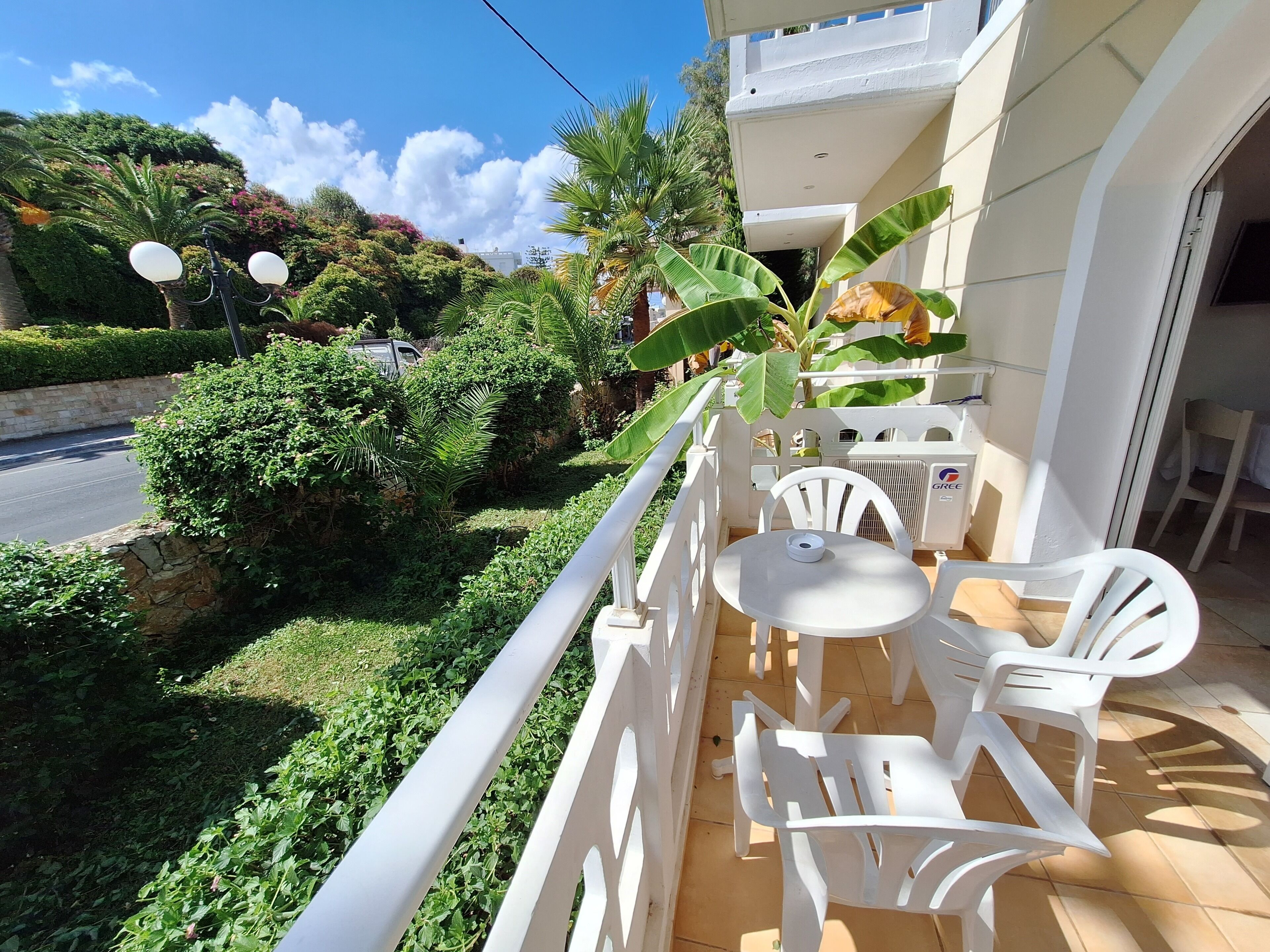 A balcony with white plastic chairs and a small round table, surrounded by lush greenery and a clear blue sky.