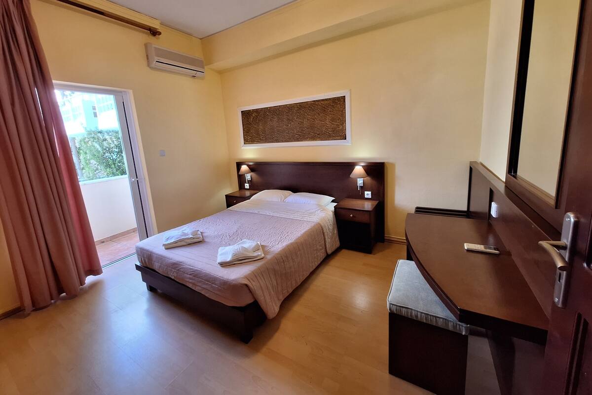 A hotel room with a bed, bedside tables, a desk, a chair, a window with curtains, and an air conditioning unit.
