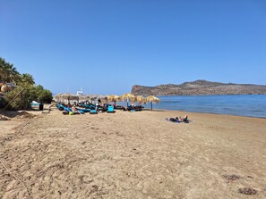 A beach with sun loungers and umbrellas, a clear sky, and a rocky shoreline. - Perla Beach (Chania)