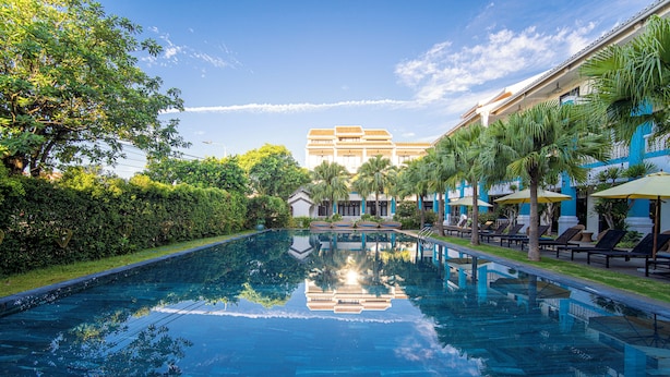 A swimming pool with a clear reflection, surrounded by palm trees and a building with a tiled roof. - Thanh Binh Riverside Hotel (Da Nang)