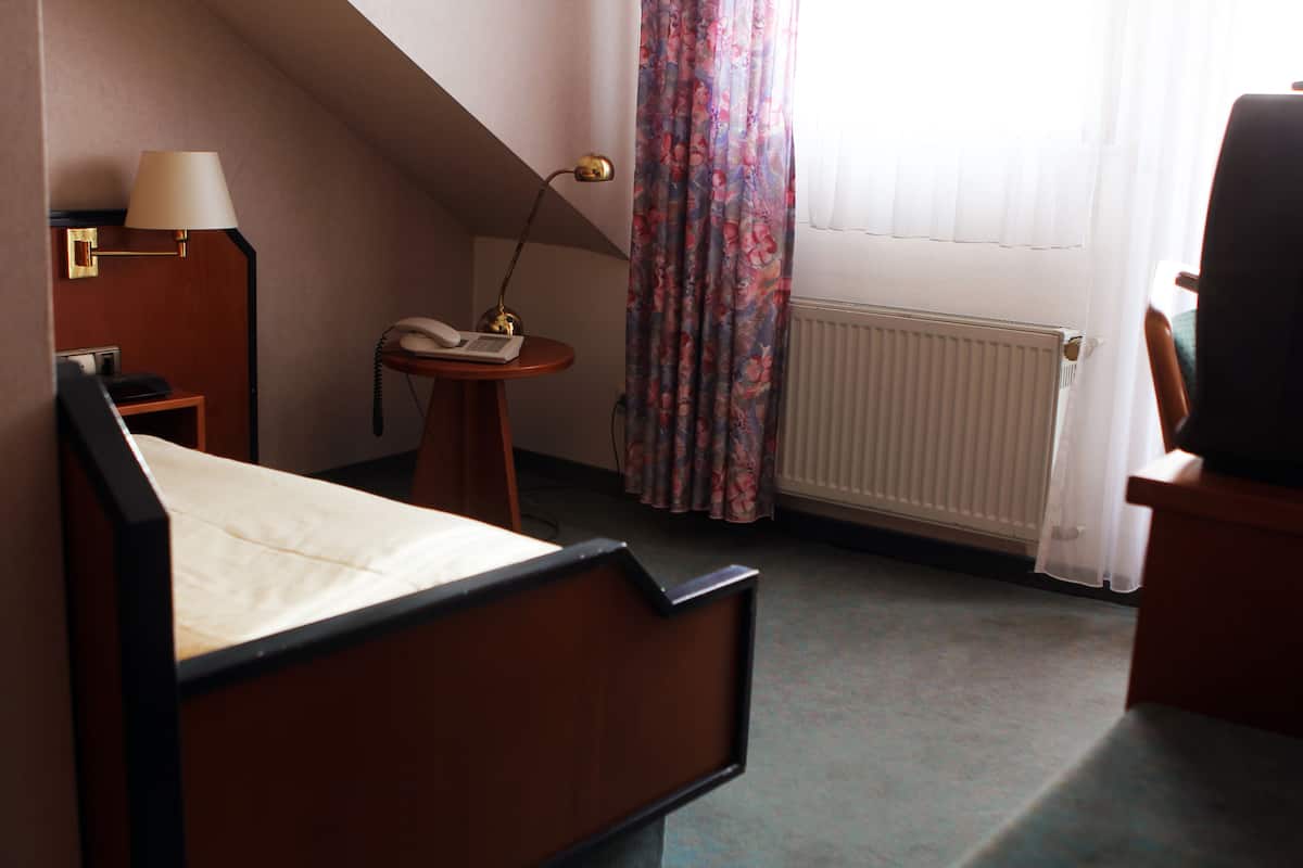 A hotel room with a bed, a desk with a lamp, a telephone, a radiator, and a window with curtains.