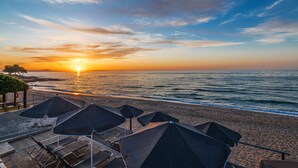 Una playa con sombrillas y sillas reclinables, con un atardecer de fondo y el océano.