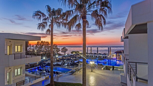 Área de piscina en un resort con sillones de descanso y sombrillas, con vista a la puesta de sol sobre el océano.