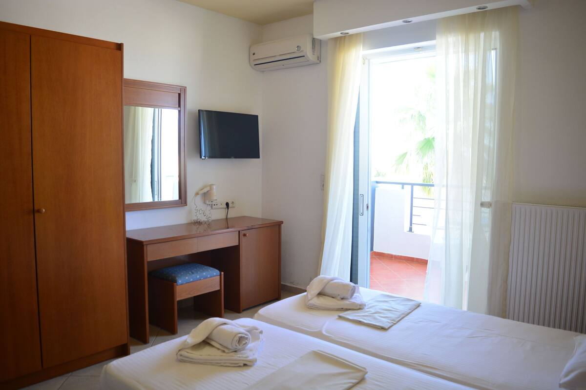A hotel room with two beds, a wooden desk, a TV, and a balcony with curtains.