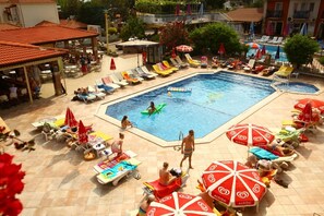 A swimming pool area with multiple sun loungers and umbrellas, surrounded by buildings with red-tiled roofs. - Turk Hotel (Fethiye)
