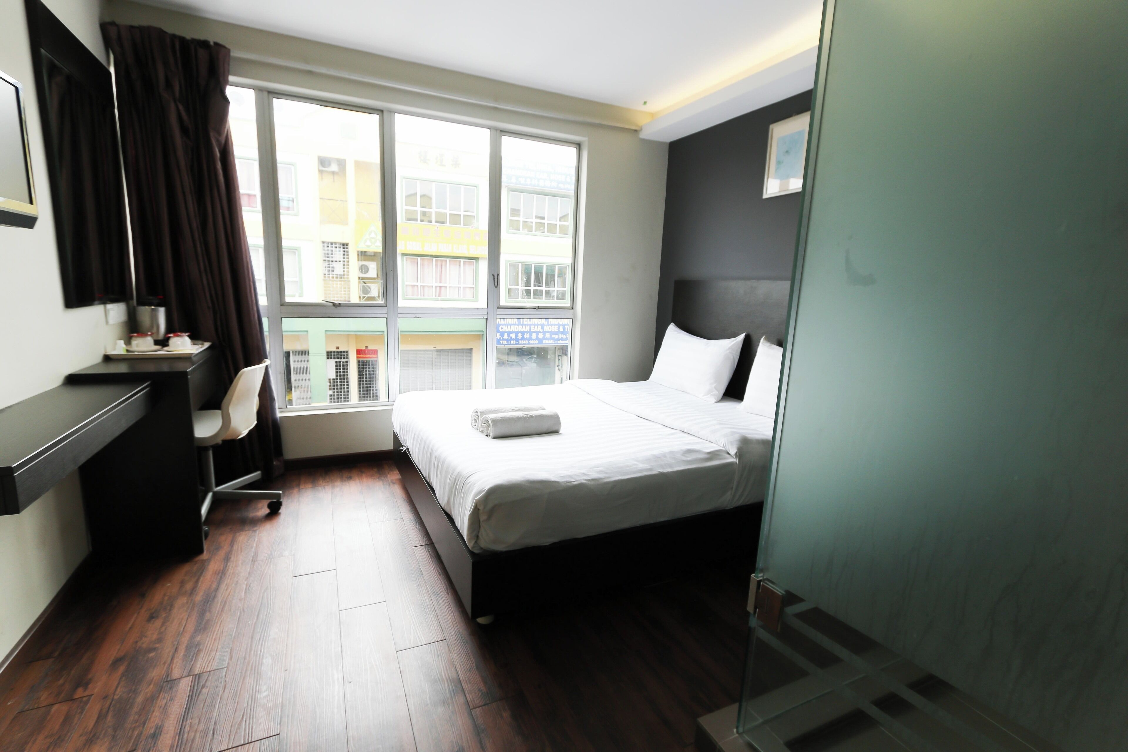 Quarto de hotel com cama, escrivaninha, cadeira e janela com vista para edifícios.