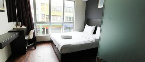 Quarto de hotel com cama, escrivaninha, cadeira e janela com vista para edifícios.