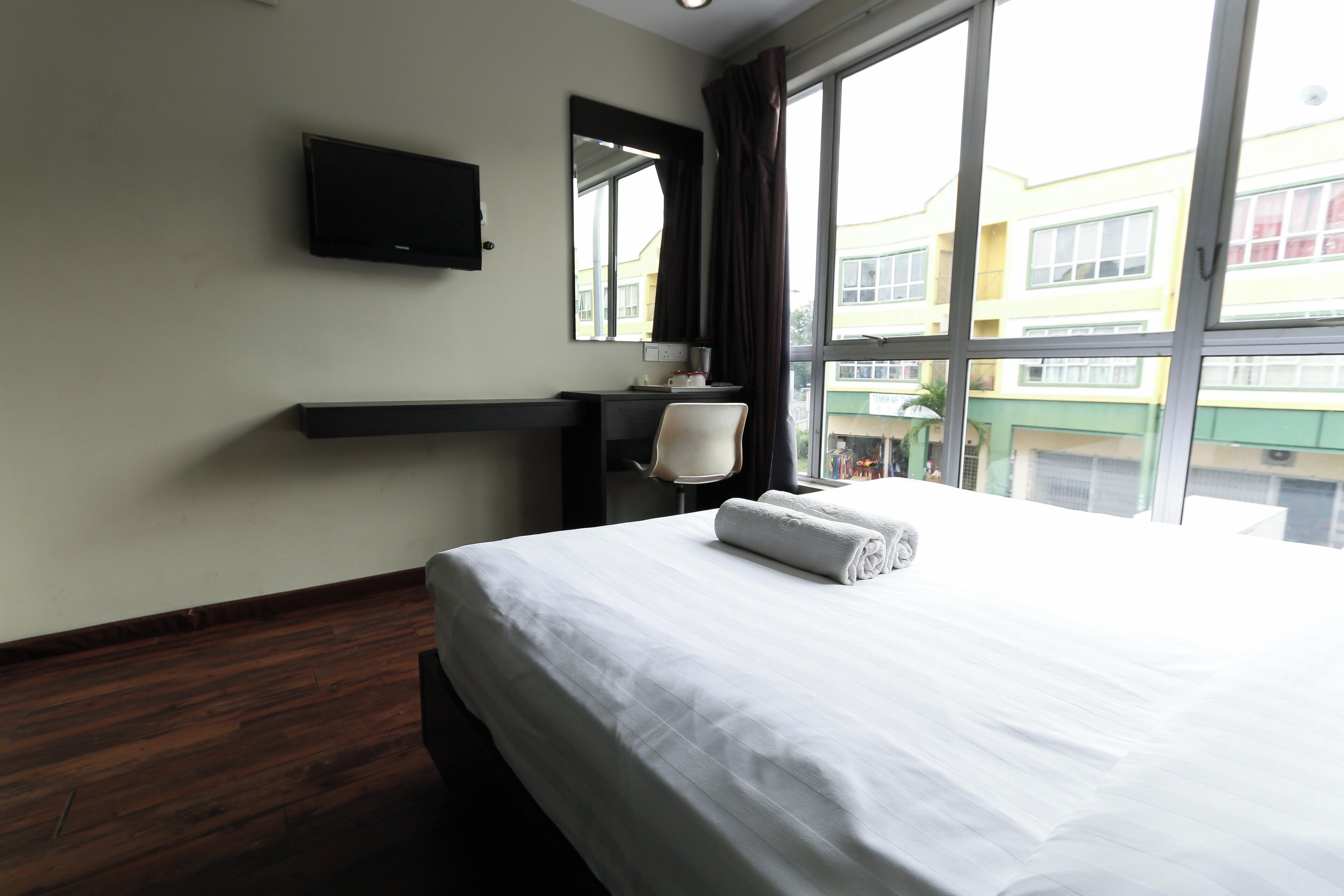 A hotel room with a bed, a flat-screen TV mounted on the wall, a desk with a chair, a window with curtains, and a view of buildings outside.