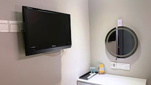 A small room with a white desk, a chair, a television mounted on the wall, and a round mirror.